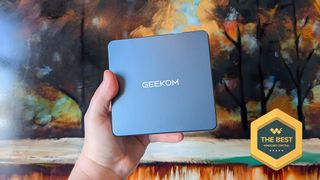 A hand holding the Geekom Mini IT13 mini PC in front of a painting of trees. A best award badge is in the bottom right corner of the image.